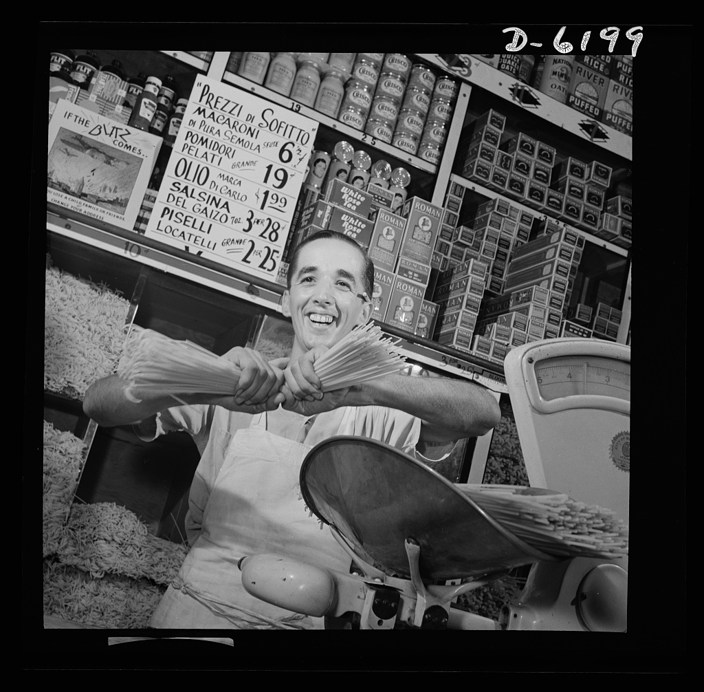A smiling Italian American grocer holding bundles of dry spaghetti beside a scale, in front of a hand-lettered price sign reading “Prezzi di Sofitto,” 1940s